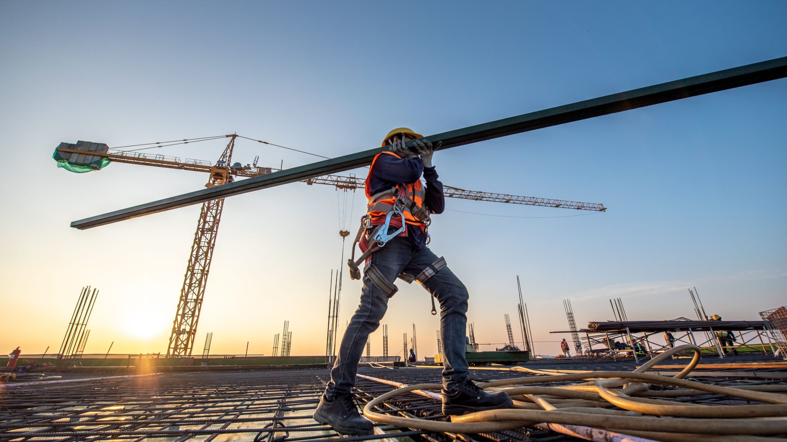 A construction worker in a safety helmet and vest carries a long metal beam across a building site at sunset. Cranes and scaffolding are visible in the background, under a clear blue sky.