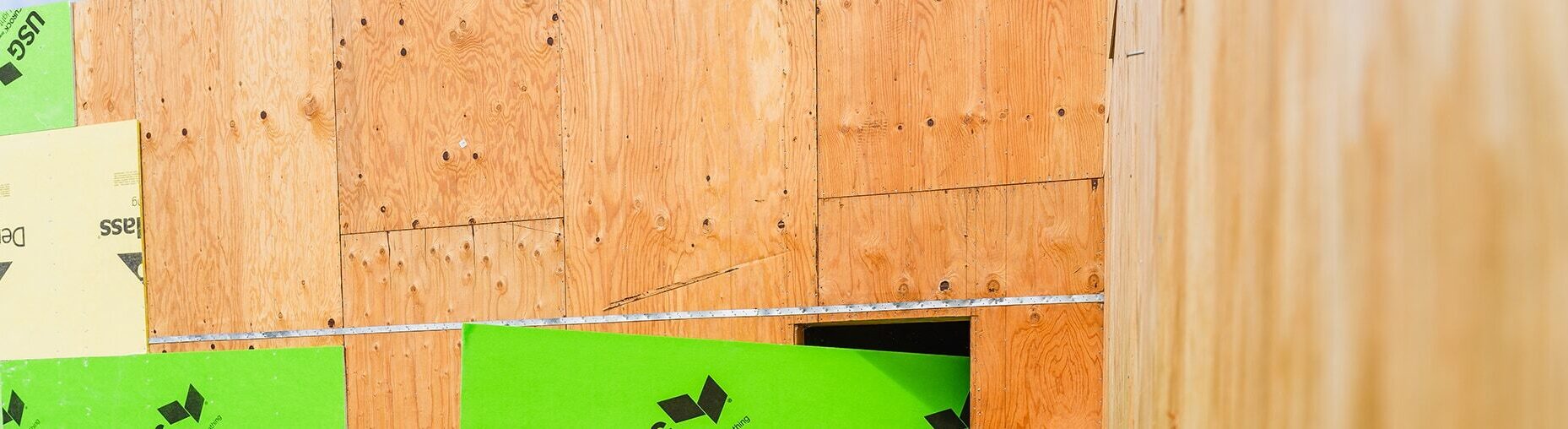 Close-up of a wooden wall under construction, with panels of plywood and sections of green sheathing material. A small rectangular opening is visible, and the wood grain is prominent on the panels.