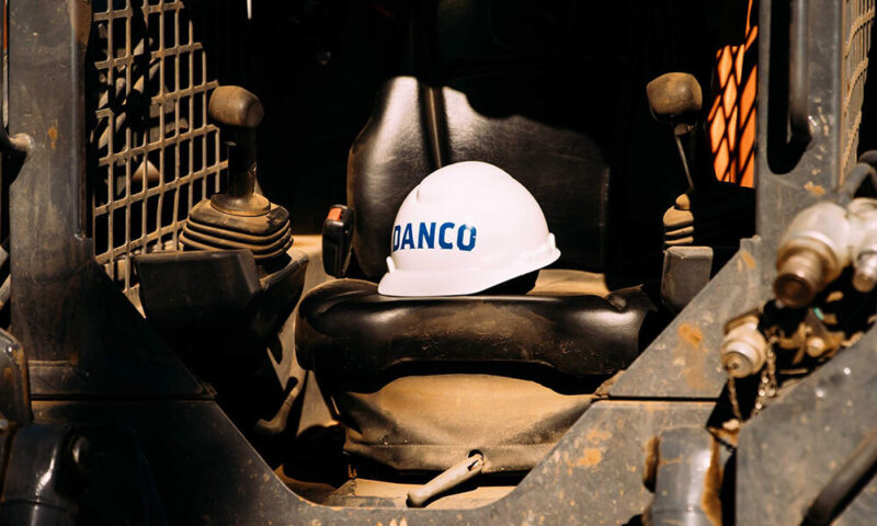 A white hard hat with DANCO written on it rests on a black seat inside the cabin of heavy machinery. The equipment controls and interior are visible, suggesting a construction or industrial setting.