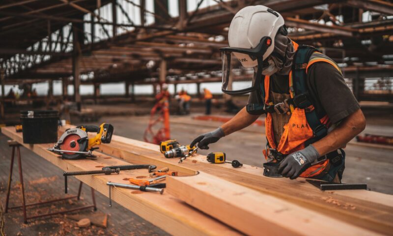 A construction worker wearing protective gear and an orange vest uses power tools to work on wooden beams at a construction site. The area is an open-frame structure with scattered tools and materials.