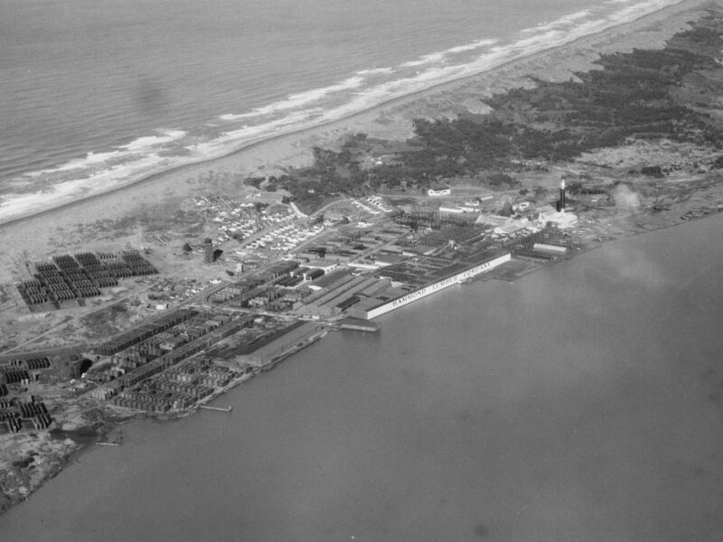 Aerial view of an industrial area on a coastline, with several buildings and structures, bordered by a sandy beach and the ocean on one side, and a body of water on the other. Vegetation is scattered throughout the landscape.