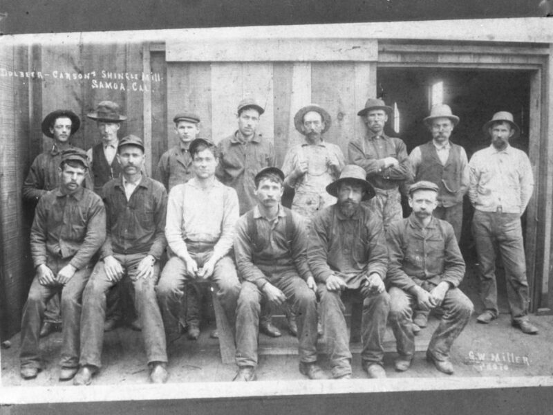 A vintage black and white photo of a group of fifteen men posing outside a wooden building. They are wearing work clothes and hats, and appear to be miners or workers. The image is labeled Driver Carson Skunk Mill, Nevada, Cal.