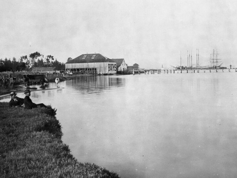A serene black-and-white photograph depicting a waterfront scene with a large building labeled Bays near the shore. Multiple ships are docked at a long pier extending into the calm water. Trees line the far bank, and a person sits on the grassy edge.