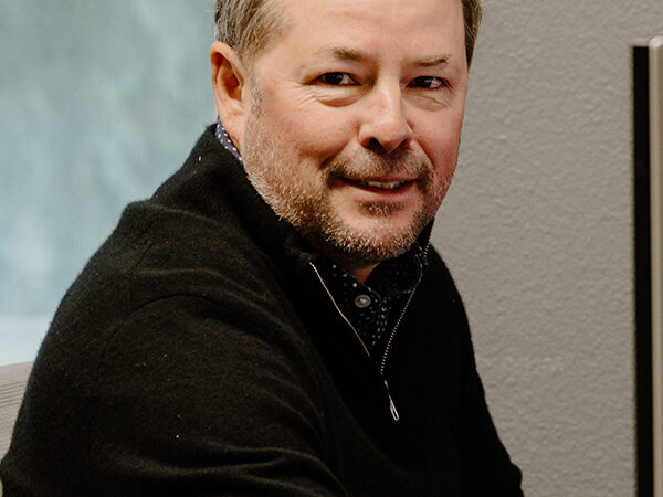 A man with short hair and a beard sits indoors, looking towards the camera. He is wearing a black sweater, and behind him is a window with a blurred view of the outdoors. The setting appears to be an office or workspace.