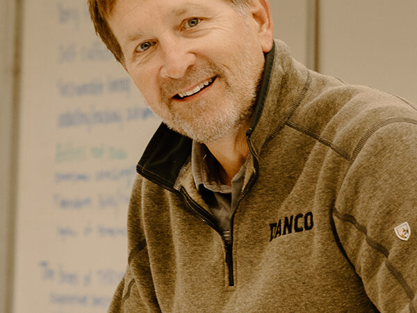 A person with a beard, wearing a gray fleece jacket with a logo, is smiling in front of a whiteboard with writing on it.