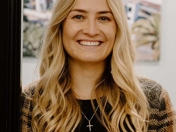 A woman with long blond hair smiles at the camera. She is wearing a plaid jacket, a black shirt, and a cross necklace. A blurred landscape painting is in the background.