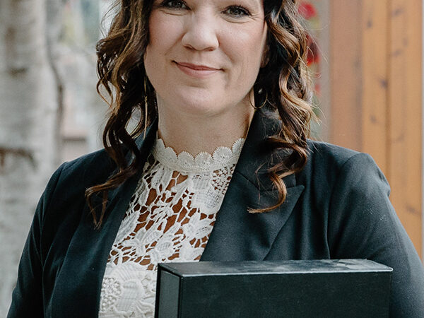 A person with curly hair, wearing a black jacket and a white lace top, holds a book and a mug. They stand outdoors in front of a wooden fence and a tree with red leaves.