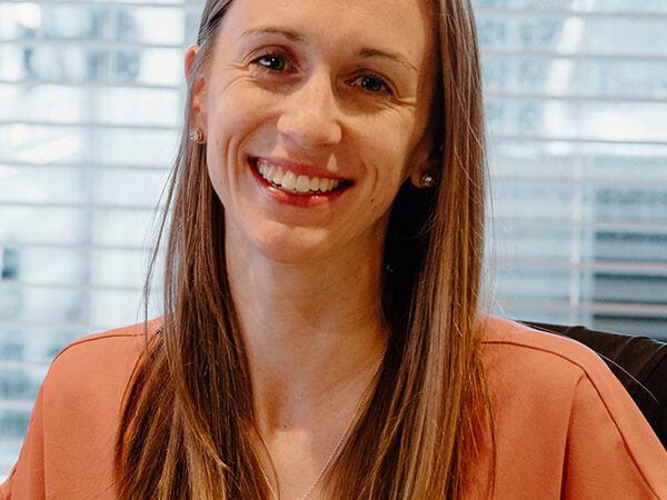 A smiling woman with long, straight, light brown hair is wearing a peach-colored top and a cross necklace. She is seated in front of a window with horizontal blinds.