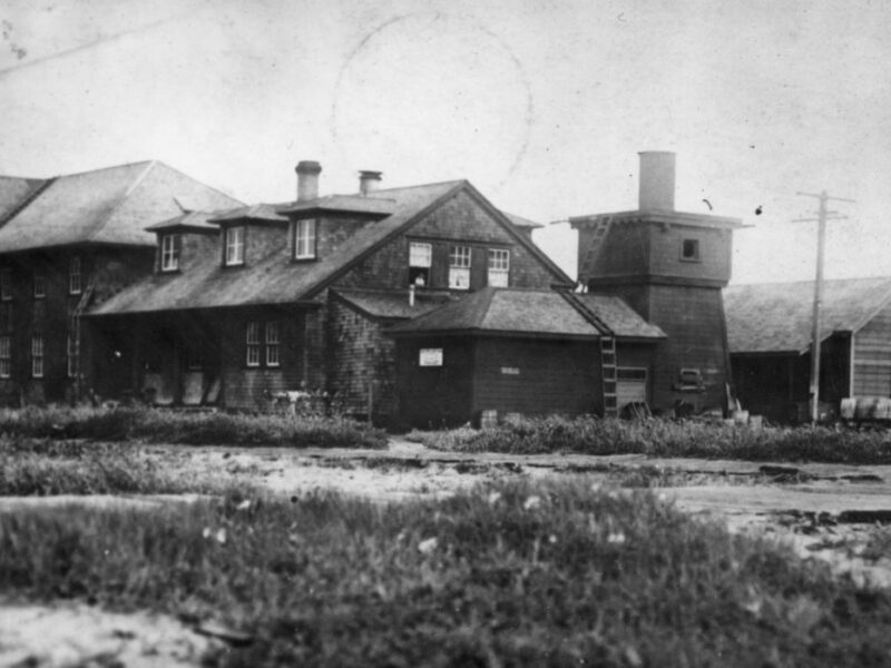A vintage black and white photo of a rustic wooden building complex with shingled roofs and multiple chimneys, surrounded by grass and sand. A small tower-like structure is attached to one of the buildings, and power lines are visible.