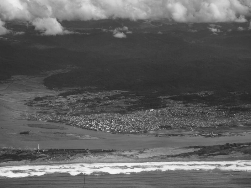Aerial view of a coastal town with dense urban areas near the shoreline. Waves crash along the sandy beach, and the background features forested hills under a partly cloudy sky. The landscape depicts a mix of natural and developed environments.