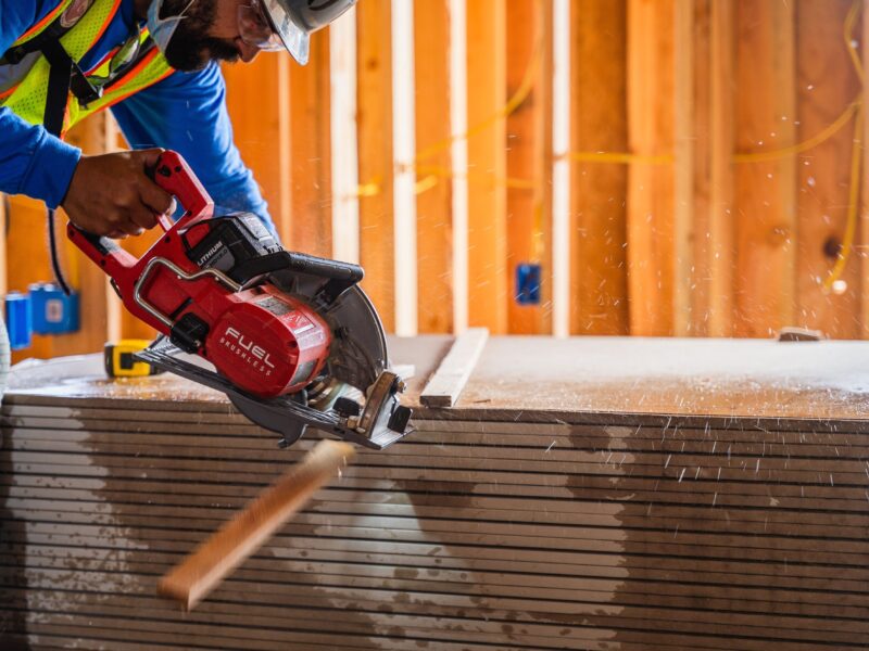 A construction worker wearing safety gear operates a red circular saw, cutting through multiple sheets of drywall. Sawdust is visible in the air. Wooden framing and electrical outlets can be seen in the background.