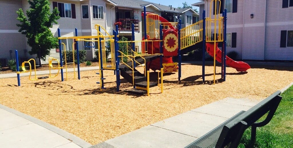 A playground with red and yellow equipment, including a slide and climbing structures, is surrounded by wood chips. Its situated in a courtyard between residential buildings, with a bench and green grass nearby.