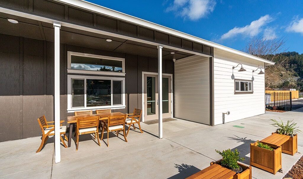 A modern building with a mix of white and brown siding, featuring a covered patio. Wooden tables and chairs are arranged outside. Planter boxes with greenery line the edge of the patio under a clear blue sky.