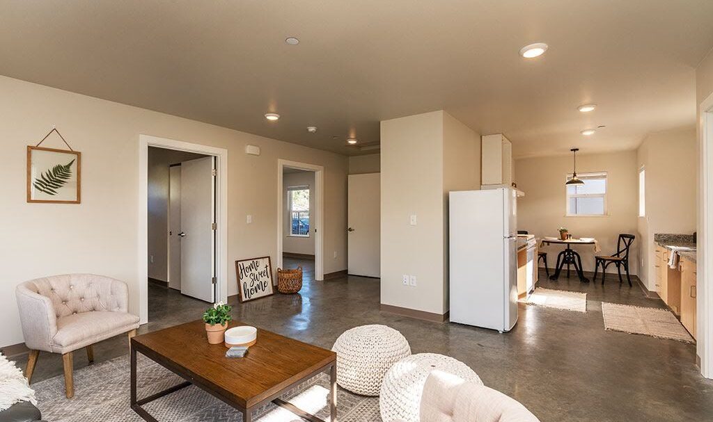 A modern apartment interior with a cozy living room featuring light beige chairs, a wooden coffee table, and poufs. In the background, theres a kitchen with white cabinets and a dining area with a table and chairs. A plant decorates the wall.