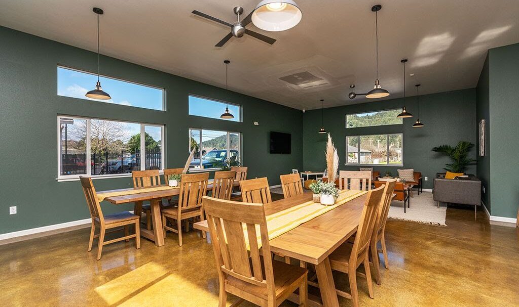 A spacious dining room with green walls and wooden furniture. Several tables and chairs are neatly arranged. Large windows provide natural light, and ceiling fans hang from above. A cozy seating area is visible in the background.