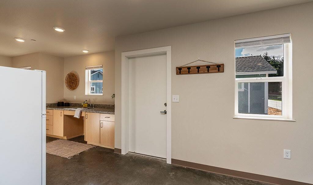 A modern kitchen with light wood cabinets, a granite countertop, and white appliances. A neutral rug lies on the floor. A window lets in natural light, and theres a decorative wall hook nearby.