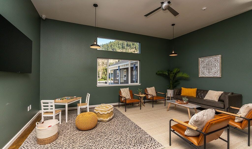 A cozy living room with green walls featuring a seating area with a sofa, armchairs, and a coffee table. A childrens play area is in the corner with small chairs and a table. A large window provides natural light. A ceiling fan and pendant lights hang above.