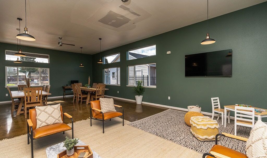 A spacious, modern living room with green walls and high ceilings. It features tan chairs, wooden tables, and pendant lighting. The room includes a kids play area with small furniture and a large wall-mounted TV. Natural light fills the space.