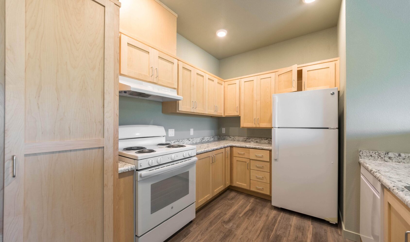 A modern kitchen with light wood cabinetry, a white oven with an overhead range hood, a white refrigerator, and granite countertops. The floor is made of dark wood, and the room is illuminated by ceiling lights.