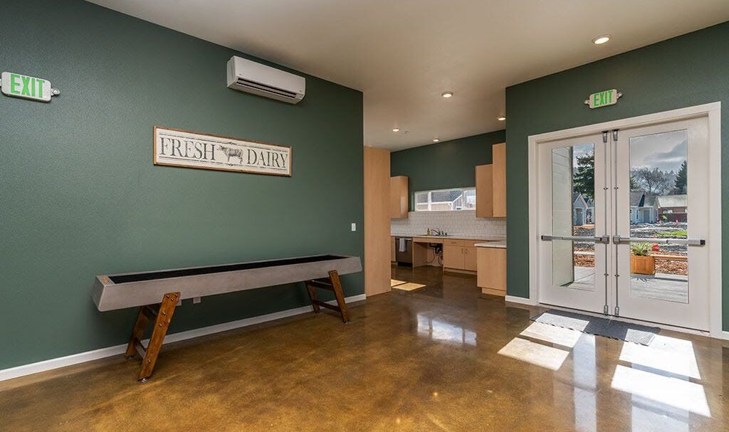 Spacious room with polished concrete floor, green walls, and a wood sign reading Fresh Dairy. A long wooden table is against one wall. The room has a small kitchenette and large glass doors leading outside. Ceiling lights illuminate the space.