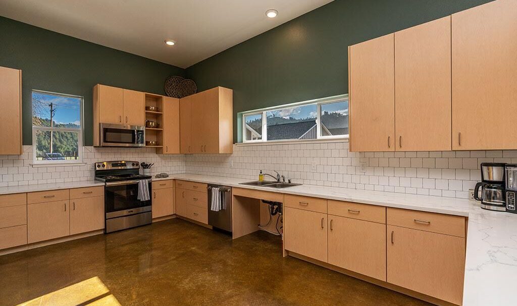 A spacious kitchen with light wood cabinets, white countertops, and a dark green accent wall. It features a stainless steel stove, oven, and microwave. The room has white tiled backsplash and natural light from windows, highlighting the polished floor.