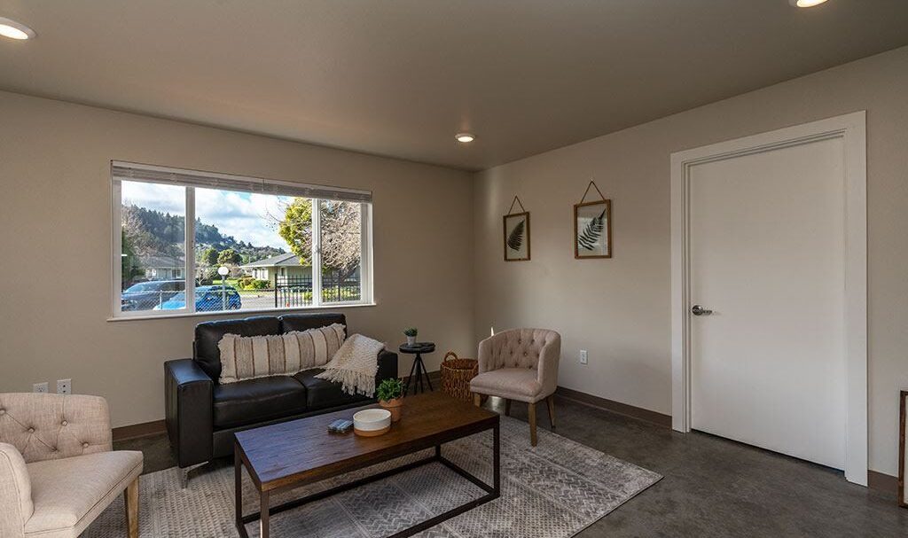 A cozy living room with a black couch and two beige chairs around a wooden coffee table. Two framed leaf prints hang on the wall. A large window lets in natural light, revealing a view of trees and a car outside.