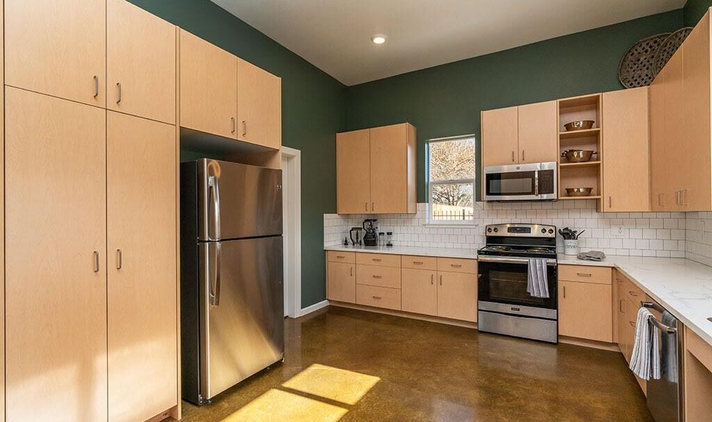 Spacious modern kitchen with light wood cabinets and stainless steel appliances, including a refrigerator, oven, and microwave. Green accent walls and white subway tile backsplash complement the polished concrete floor. Sunlight illuminates the room.