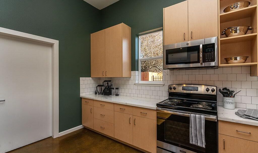 A modern kitchen with green walls and wooden cabinets. It features an electric stove, microwave, and a white tiled backsplash. A window lets in natural light, and a blender is on the counter beside a towel hanging on the oven handle.