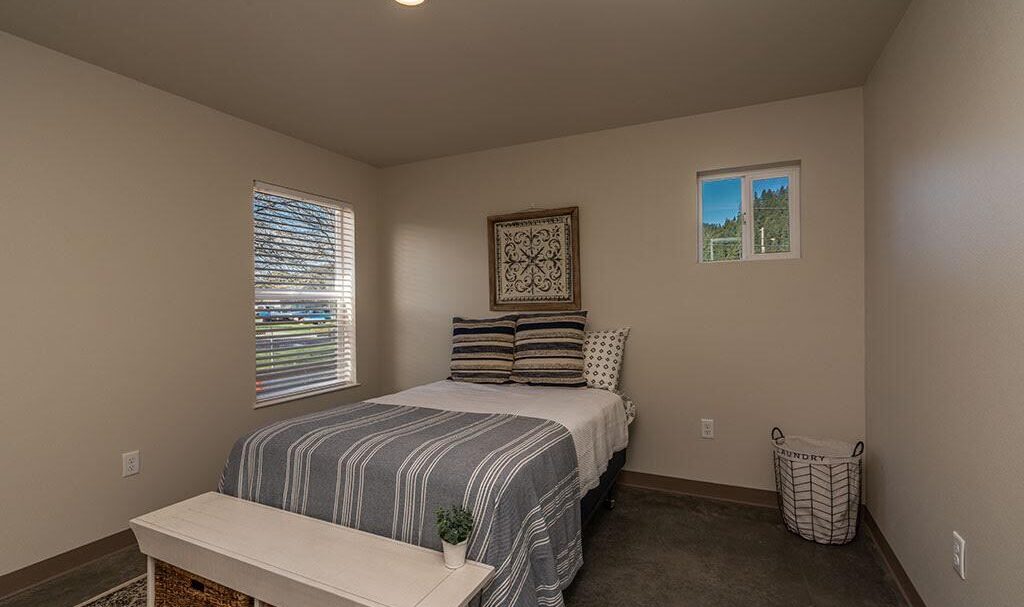 A small bedroom with a neatly made bed featuring striped bedding and multiple pillows. A decorative wall piece hangs above the bed. A bench is placed at the foot of the bed, and a window lets in natural light. A laundry basket is in the corner.