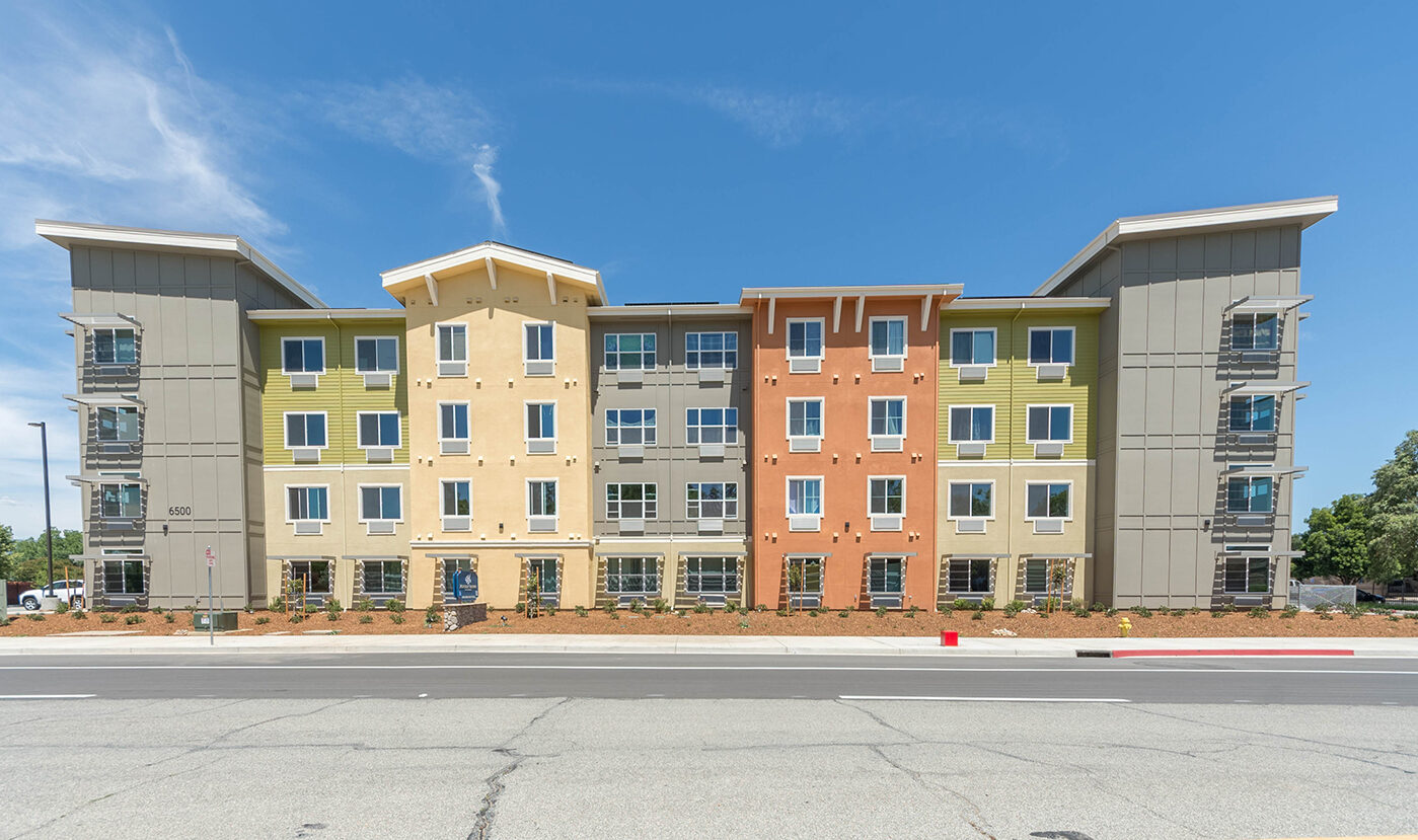 Modern four-story apartment building with a mix of yellow, orange, and green facade panels, large windows, and a sloped roof. The surrounding landscape includes young shrubs, and a wide road is visible in the foreground under a clear blue sky.