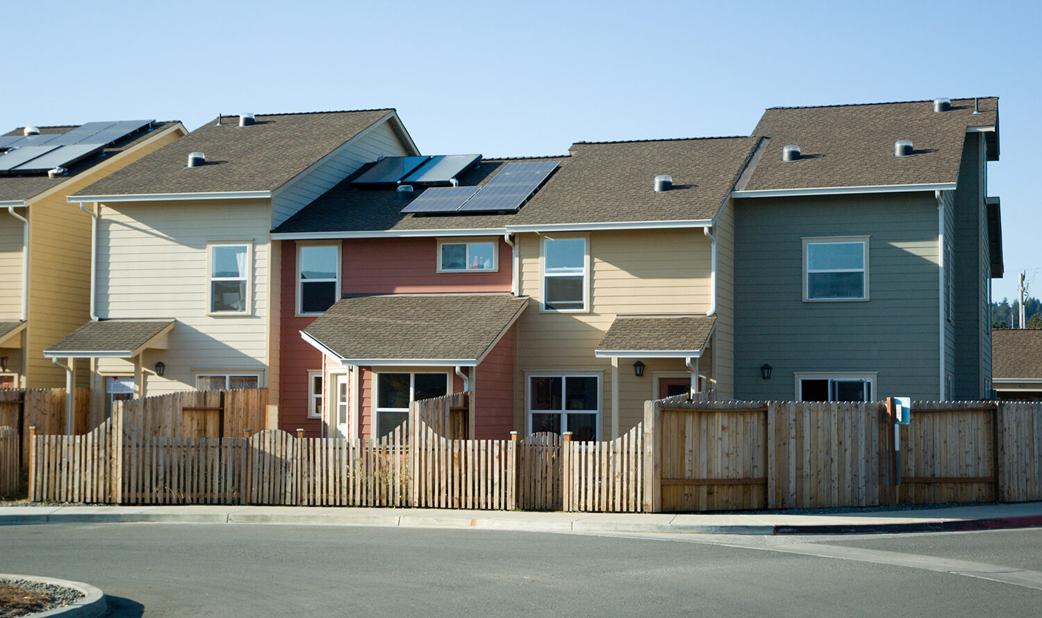 A row of modern, two-story townhouses with slanted roofs and solar panels. The houses are painted in pastel yellow, pink, and beige. Wooden fences separate each property, and the sky is clear and blue.