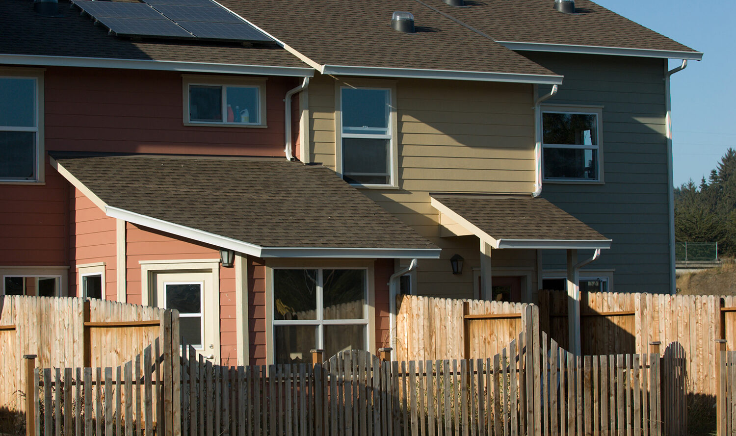 A row of colorful townhouses with solar panels on the roof, featuring red, yellow, and blue exteriors. A wooden fence runs along the front, and trees are visible in the background under a clear blue sky.
