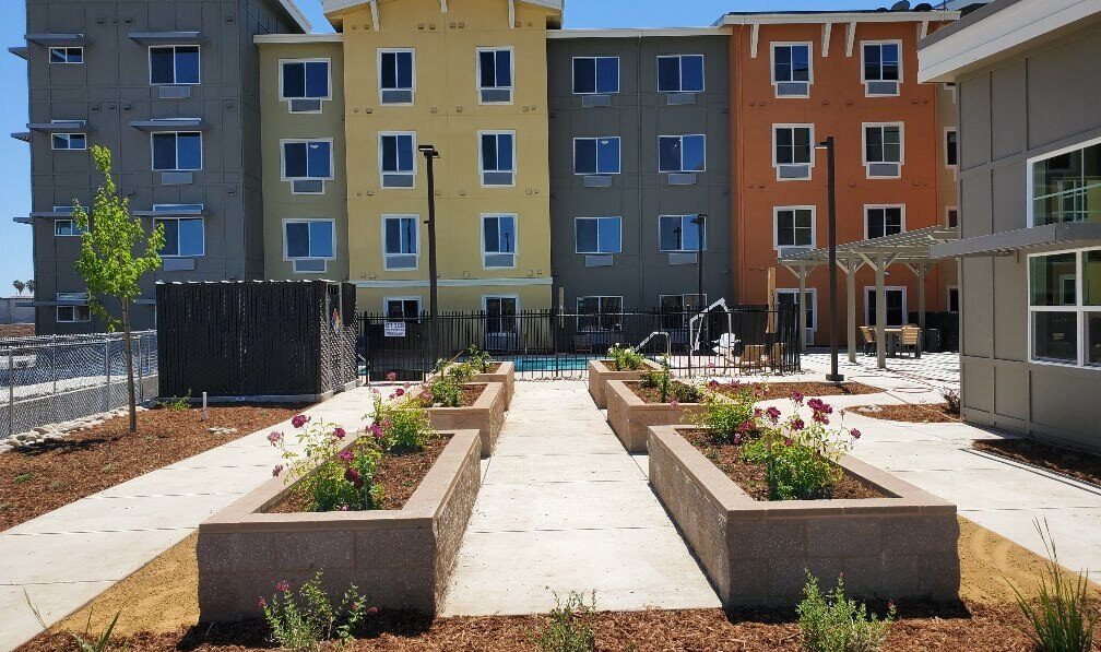A view of an apartment complex with four stories, featuring earth-toned colors. In the foreground, there are raised garden beds with blooming flowers lining a paved pathway. A fenced pool area is visible in the middle background. Clear blue sky above.