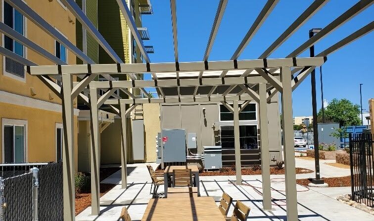 Outdoor area with a pergola casting striped shadows on concrete. Wooden tables and chairs are arranged in a line, surrounded by a yellow building, greenery, and a clear blue sky.