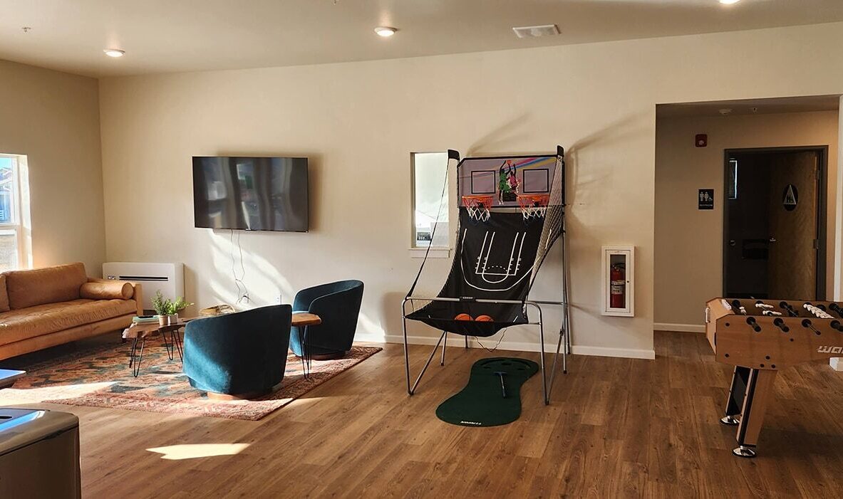 A cozy recreation room with a couch and two armchairs facing a TV on the wall. Theres a basketball arcade game and a foosball table on the right. The floor is wooden, and sunlight streams through a window on the left.