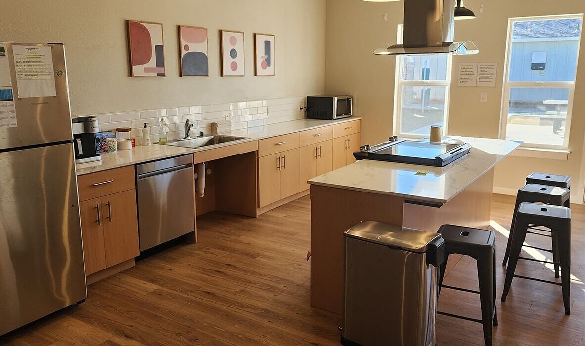 A modern kitchen with wooden floors and cabinets. Features include a stainless steel fridge, dishwasher, microwave, and an island with bar stools. Artwork decorates the wall above the sink. Large windows provide natural light.
