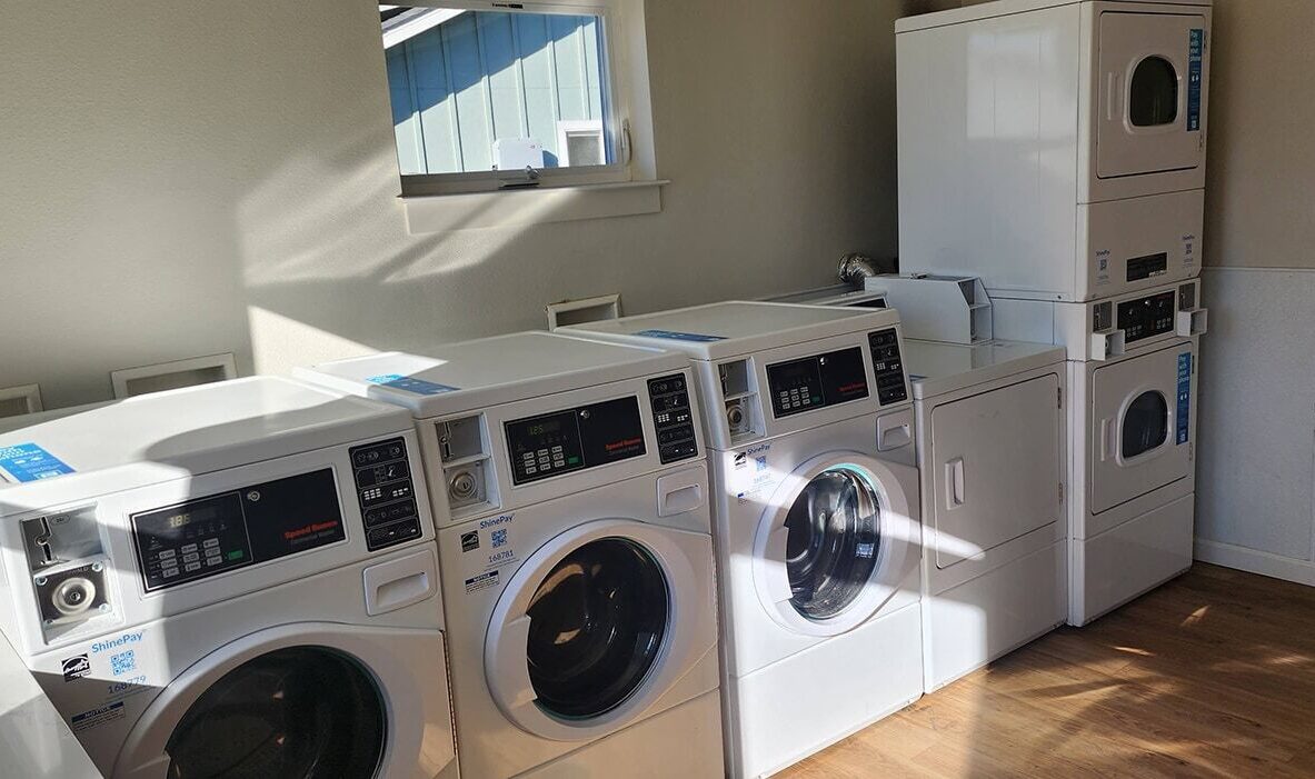 A laundromat with three front-loading washing machines in a row, followed by two stacked dryers against a beige wall. Sunlight streams in through a small window, casting shadows on the wooden floor.