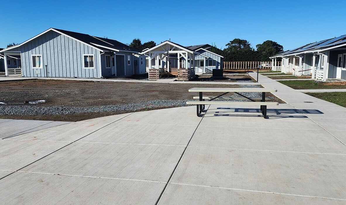 A cluster of small, single-story houses with light blue siding and white trim. The area features tidy pathways and a picnic table on a concrete surface under a clear blue sky. Solar panels are visible on some rooftops.