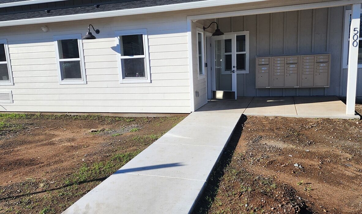 A single-story building with a white exterior and a small covered entrance. It has several windows and a row of mailboxes mounted on the wall near the door. A concrete path leads from the foreground to the entrance over a patch of bare earth.