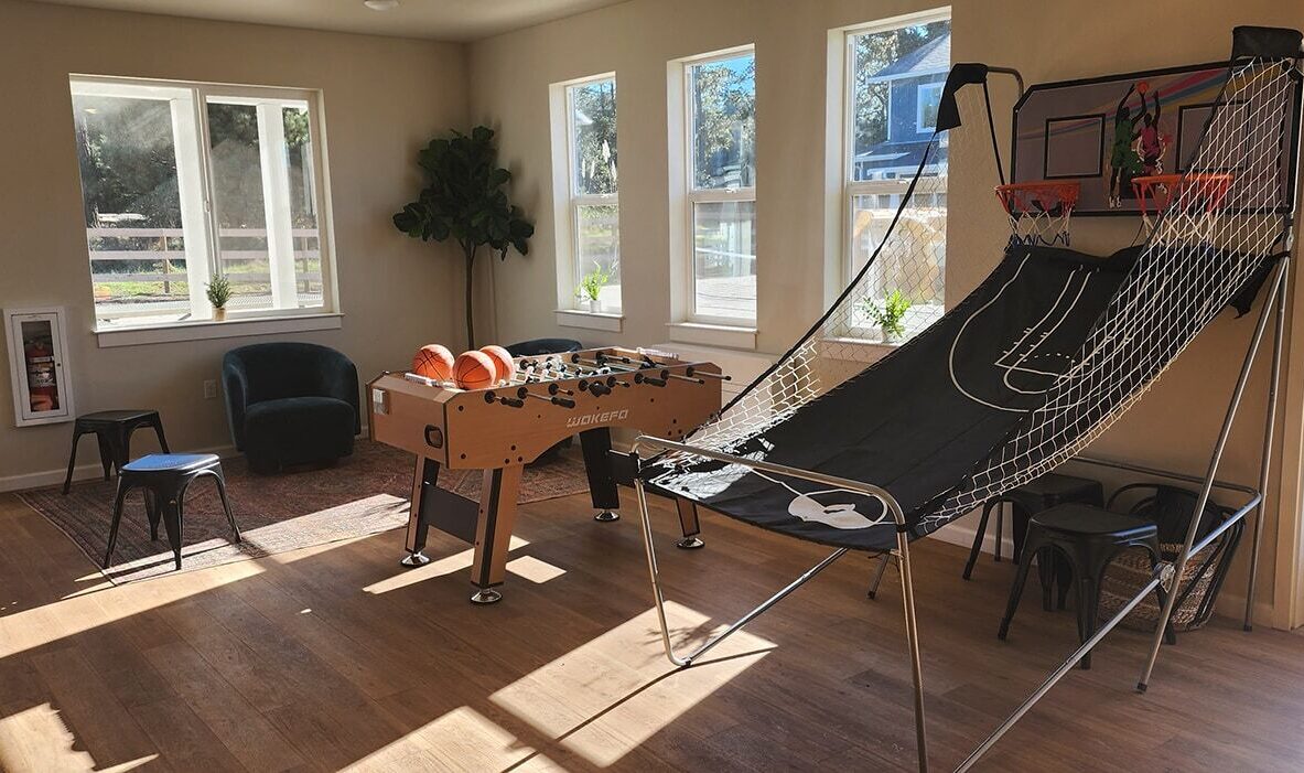 A sunlit recreation room with a foosball table and an arcade basketball game. The room has wooden floors, several chairs, a potted plant, and large windows letting in natural light.
