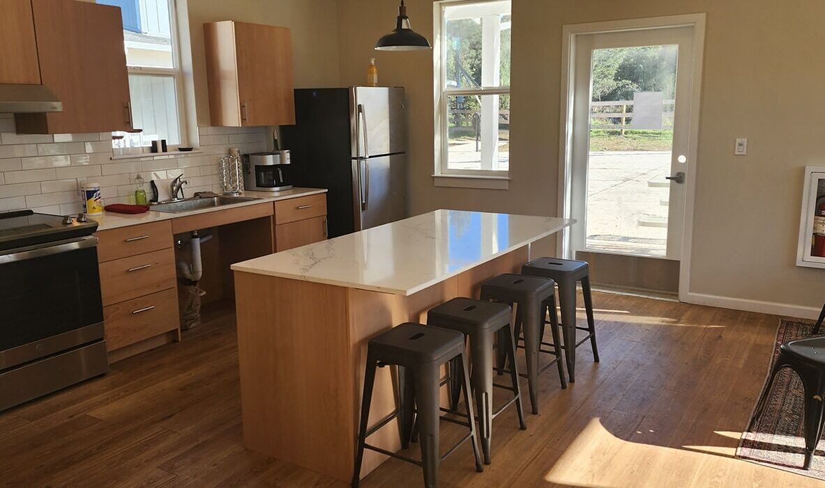 A modern kitchen with wooden cabinets, a stainless steel refrigerator, and a black oven. A white marble island has four black stools. Large windows provide natural light. A fire extinguisher is mounted on the wall. A single black chair is in the corner.