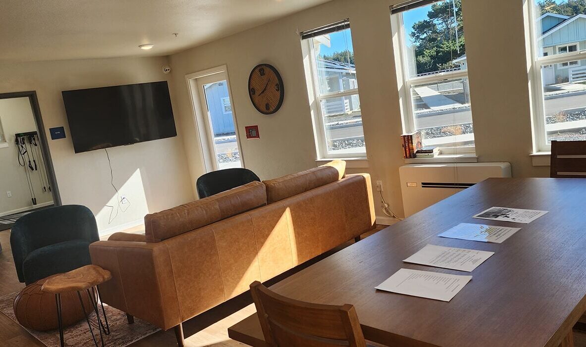 A cozy living room with a brown leather sofa, wooden coffee table, and TV mounted on the wall. Large windows let in natural light. A wooden dining table with papers is in the foreground. A pouf and two cushioned chairs complete the seating arrangement.