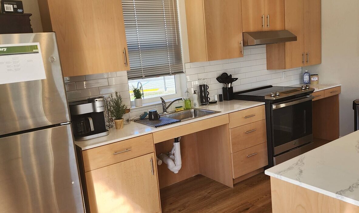 A modern kitchen with light wood cabinets, a stainless steel refrigerator, stove, and oven. A white countertop and a small sink are visible. A coffee maker and dish rack are on the counter, with a striped window blind above the sink.