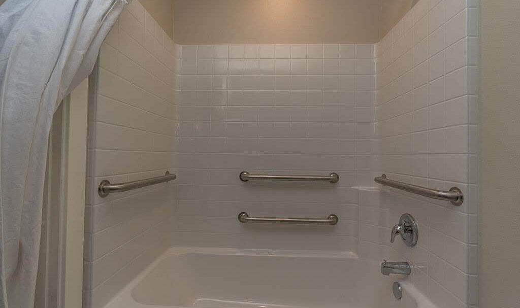 A bathroom with a white tiled shower and bathtub, featuring three metal grab bars on the walls. A white shower curtain is partially visible on the left. The faucet and showerhead are on the right.
