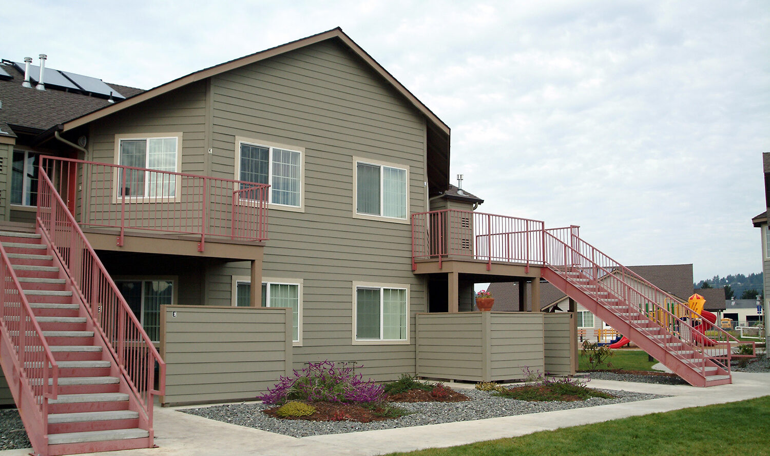 Two-story apartment building with beige siding and red stair railings. Stairs lead to upper apartments. Landscaping includes grass and small shrubs. Playground visible in the background. Overcast sky.