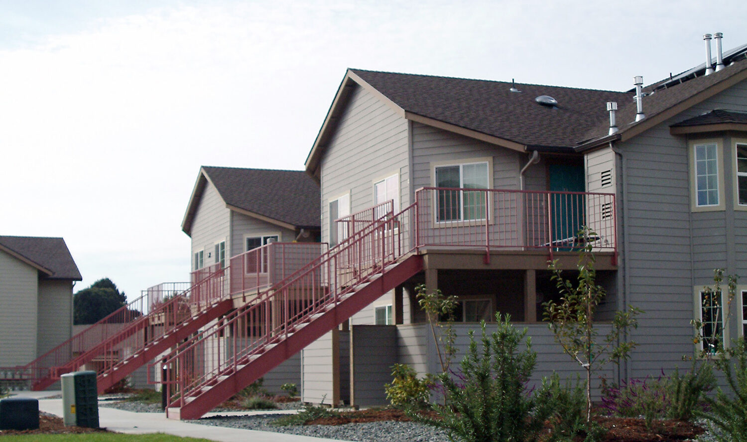 Apartment buildings featuring beige siding and red staircases under a partly cloudy sky. The foreground has a curved pathway lined with mulch and shrubs, leading through a landscaped area.