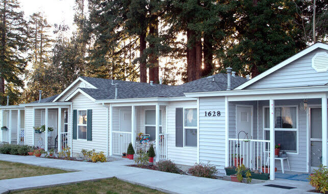 A row of small, white single-story houses with porches, surrounded by tall trees. Each house has a light blue door and potted plants on the porch. The sun is setting, casting a warm glow on the scene. House number 1628 is visible.