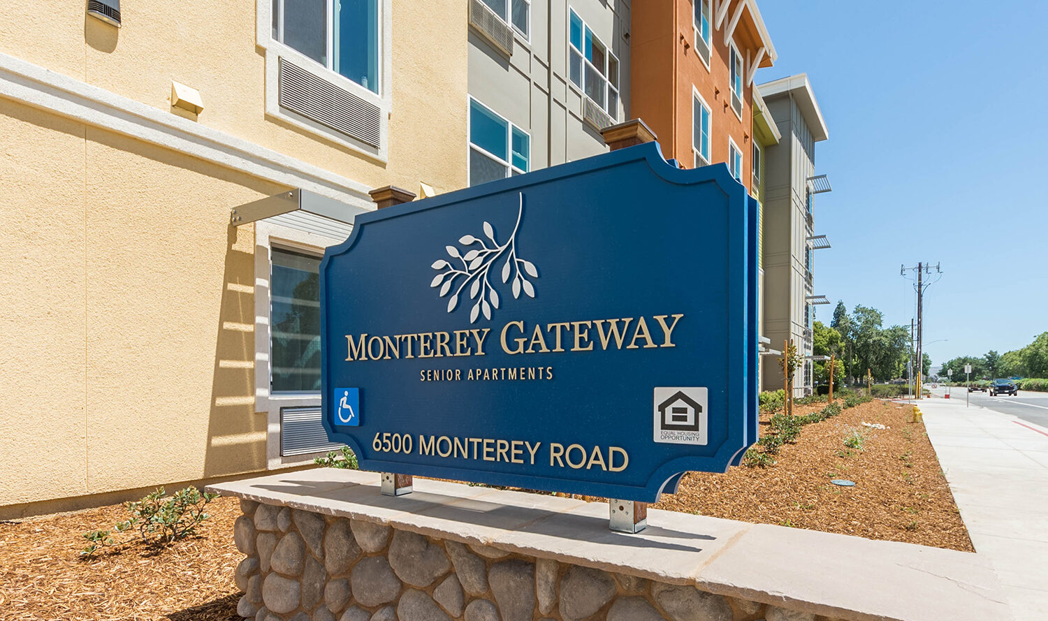 Sign for Monterey Gateway Senior Apartments in front of a modern apartment building. The sign includes accessibility and equal housing opportunity symbols. The building is surrounded by landscaped grounds and a clear blue sky is in the background.