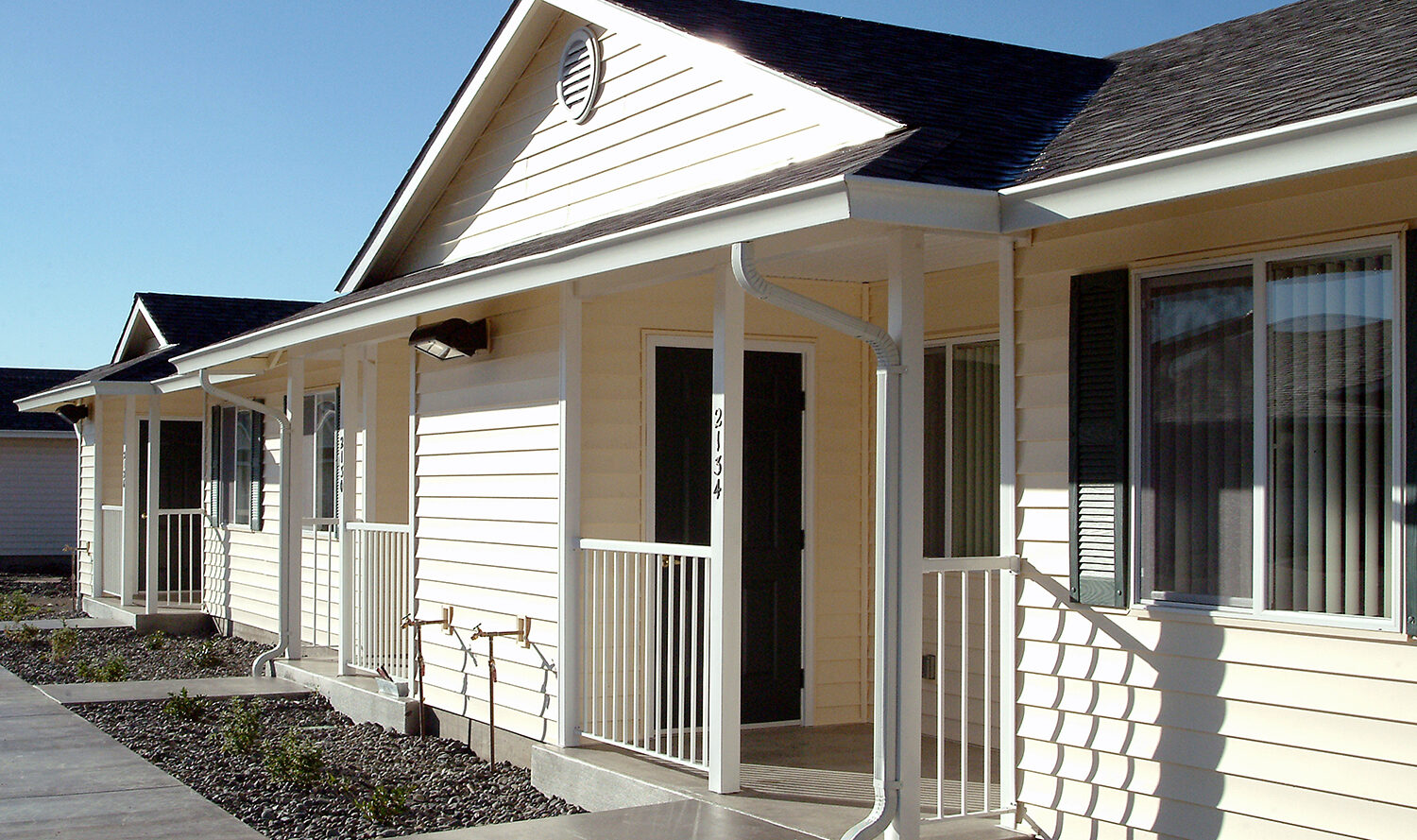A row of beige single-story houses with dark roofs and white trim, featuring small front porches and gravel landscaping. A clear blue sky is in the background.
