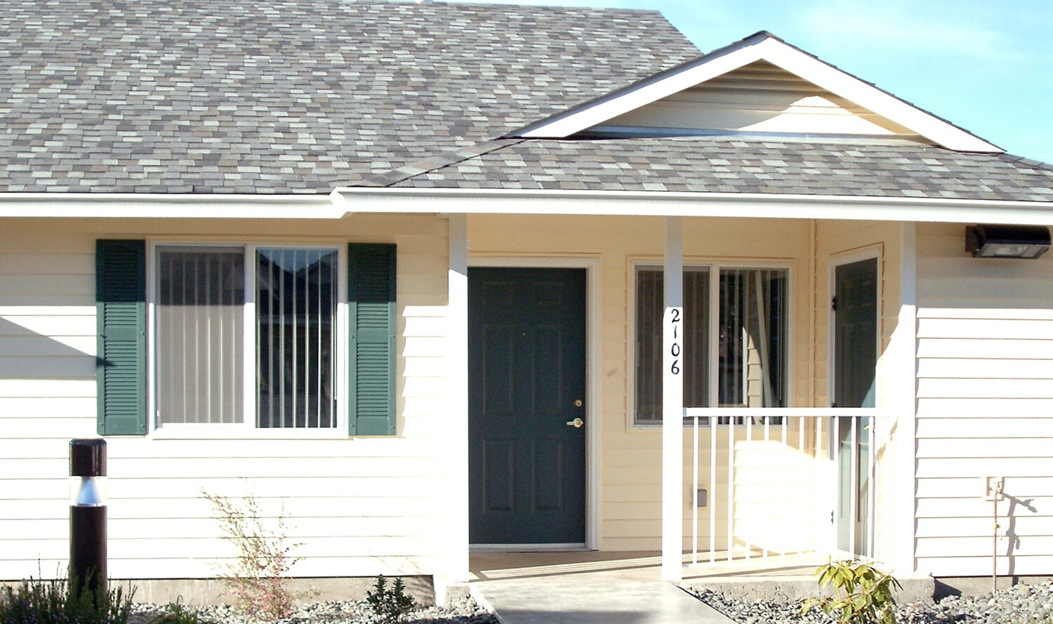 A one-story house with a gray shingle roof, white siding, and green shutters. There is a blue front door surrounded by windows. A path leads to the entrance, with small shrubs lining the walkway. The sky is clear and blue.